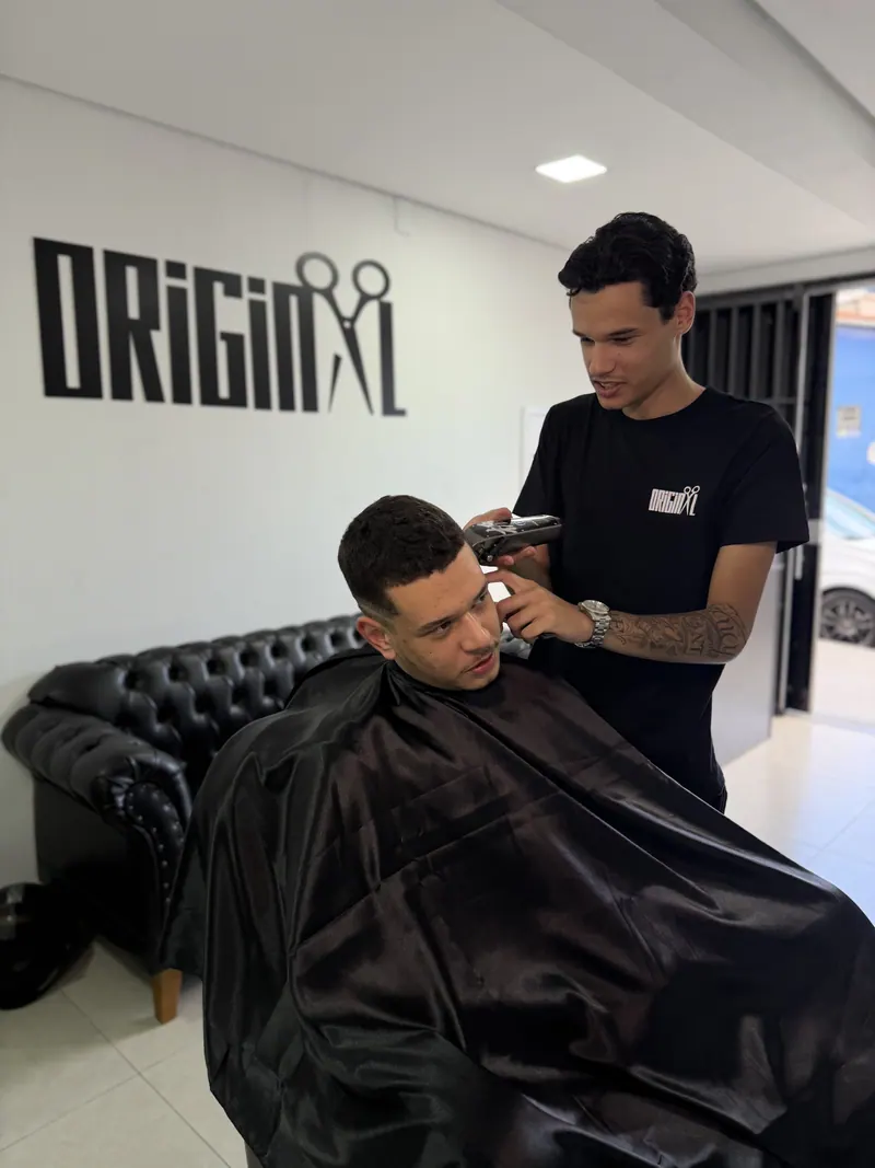 Barbeiro da Original Barbershop finalizando um corte com a logo da barbearia ao fundo